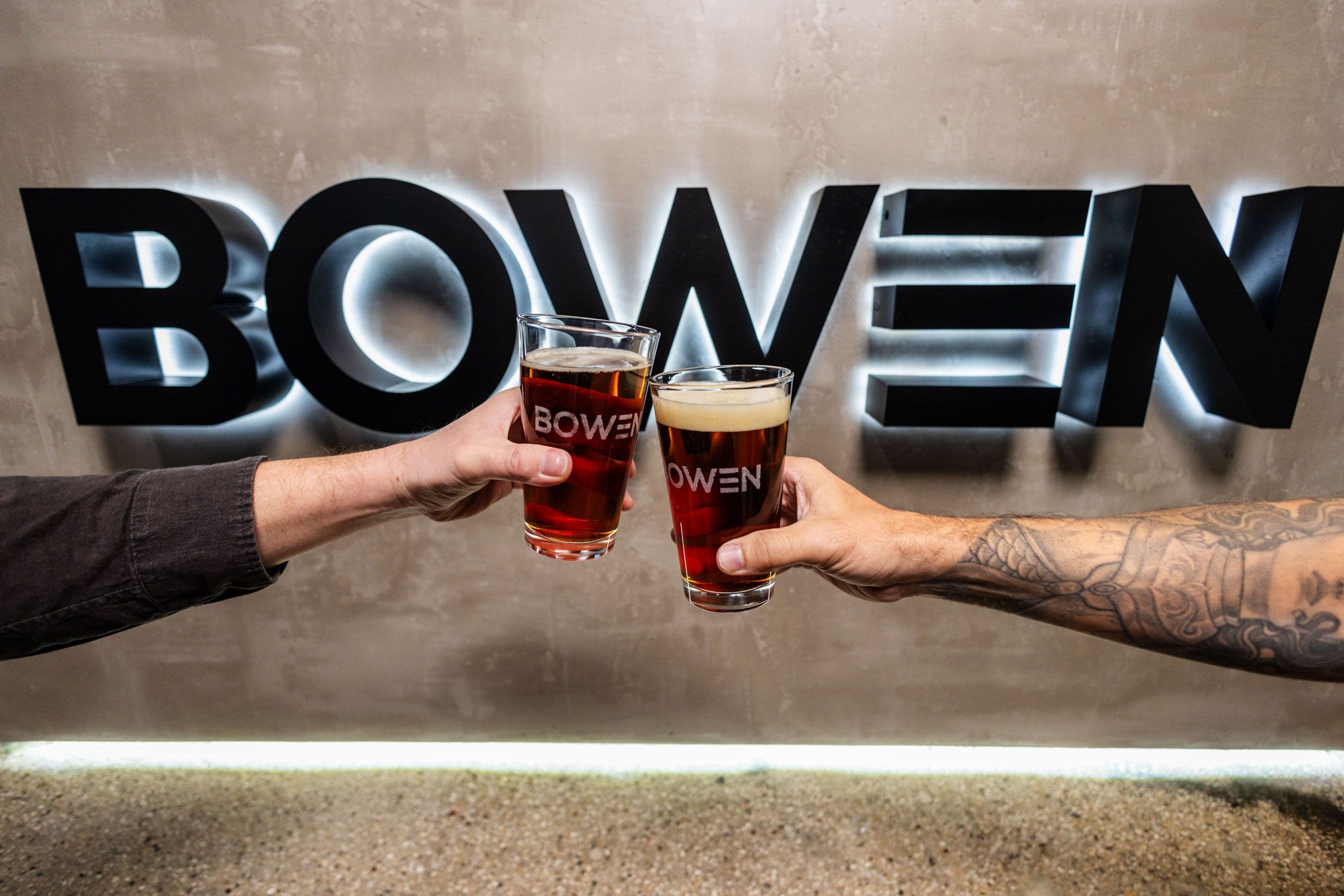 Two individuals cheerfully hold up beer glasses in front of a sign that reads "BOWEN"